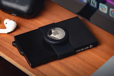 Slim black wallet with AirTag strap attached, placed on a wooden desk beside tech accessories.